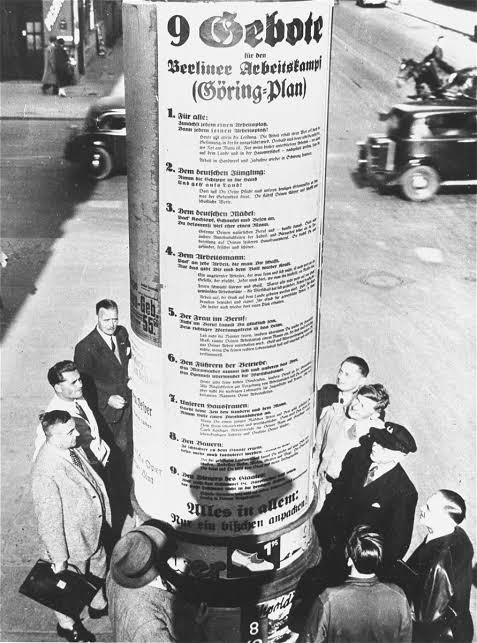 Men reading Nazi propaganda pasted to a large pillar.